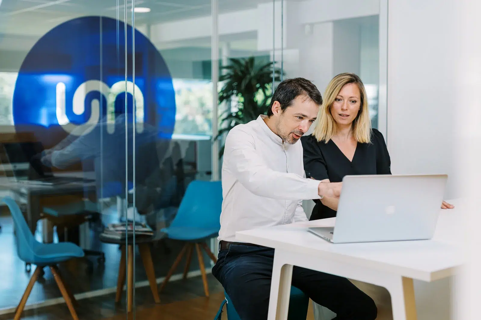 Deux collègues discutent devant un ordinateur portable dans un bureau moderne vitré.
