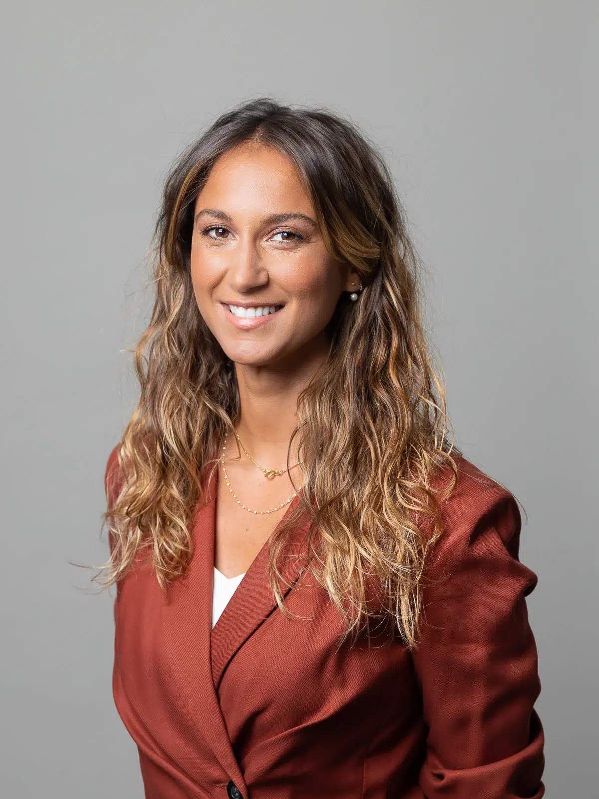 Portrait professionnel d'une femme souriante en veste rouille sur fond gris.