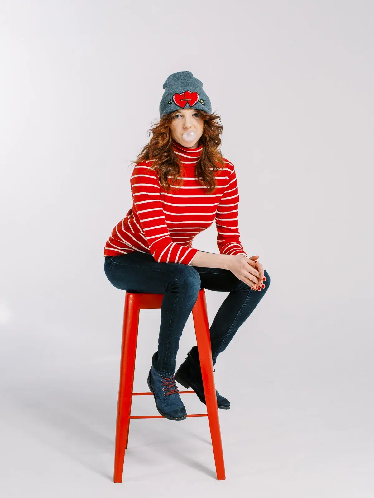 Jeune femme assise sur un tabouret rouge, portant un bonnet et faisant une bulle de chewing-gum.