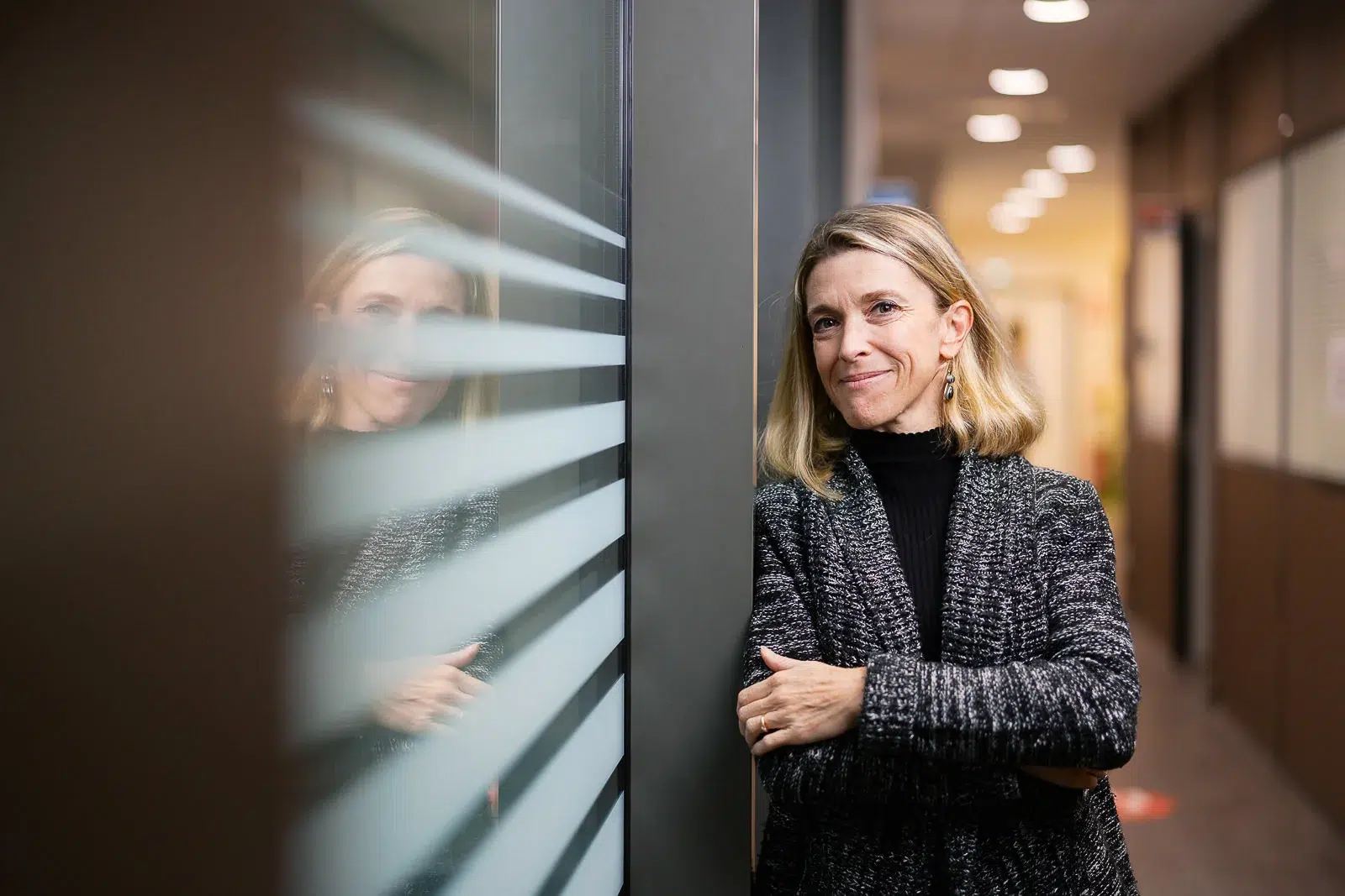 Femme souriante adossée à une paroi vitrée dans un couloir de bureau moderne.