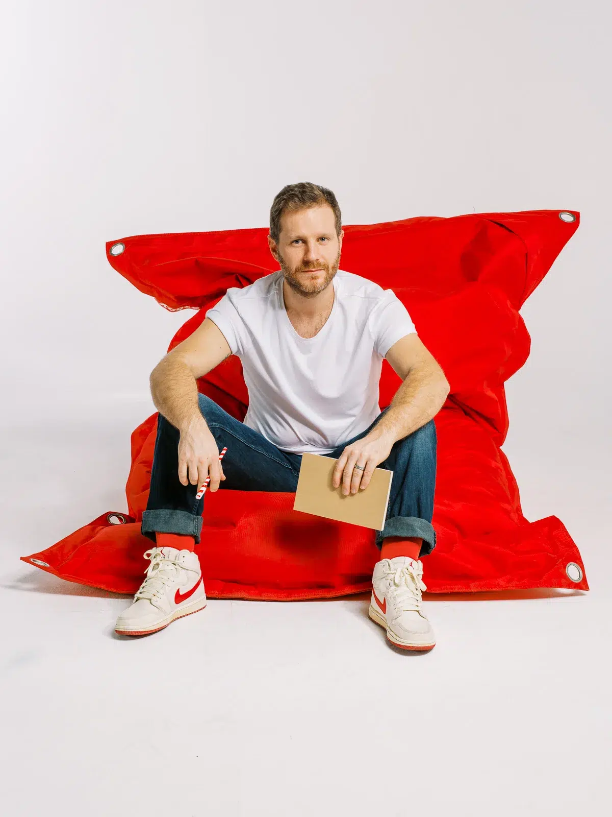 Homme assis sur un grand pouf rouge, tenant un carnet et un stylo, sur fond blanc.