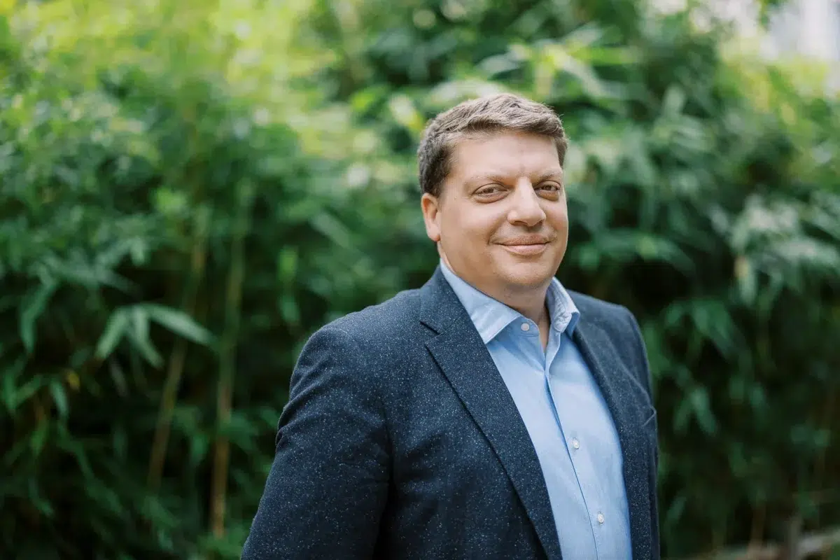Portrait d'un homme souriant en veste bleue et chemise claire, avec un fond de feuillage vert.