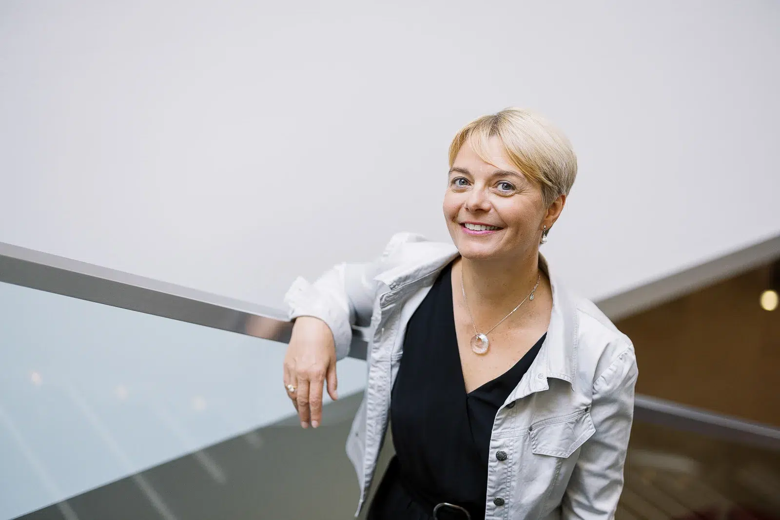 Femme souriante en veste claire adossée à la rambarde d'un escalier moderne.