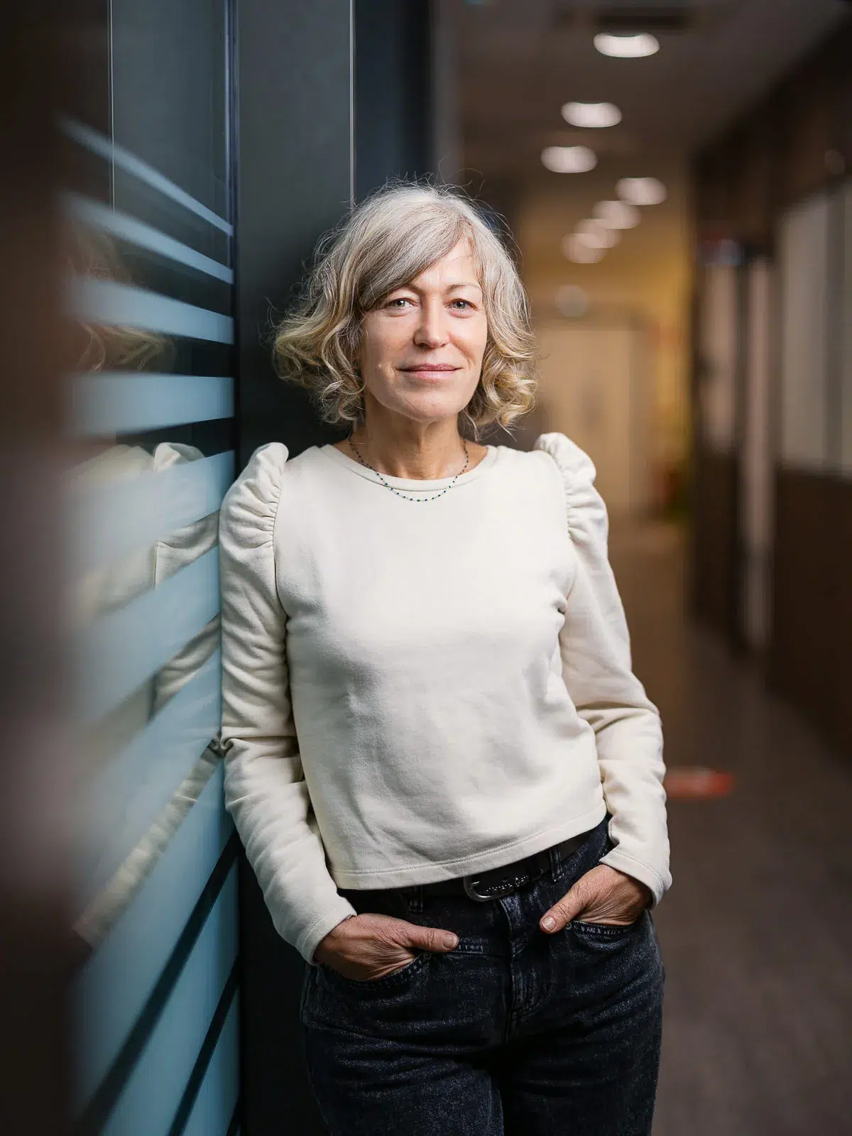 Femme souriante en pull clair et jean, les mains dans les poches, dans un couloir de bureau.