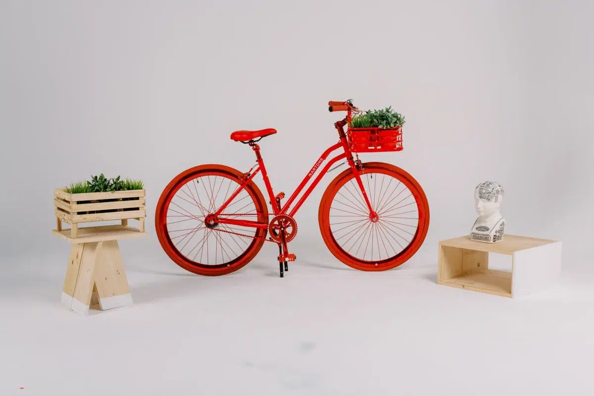Vélo rouge vif avec panier fleuri, entouré de petits meubles en bois et d'un buste décoratif sur fond blanc.