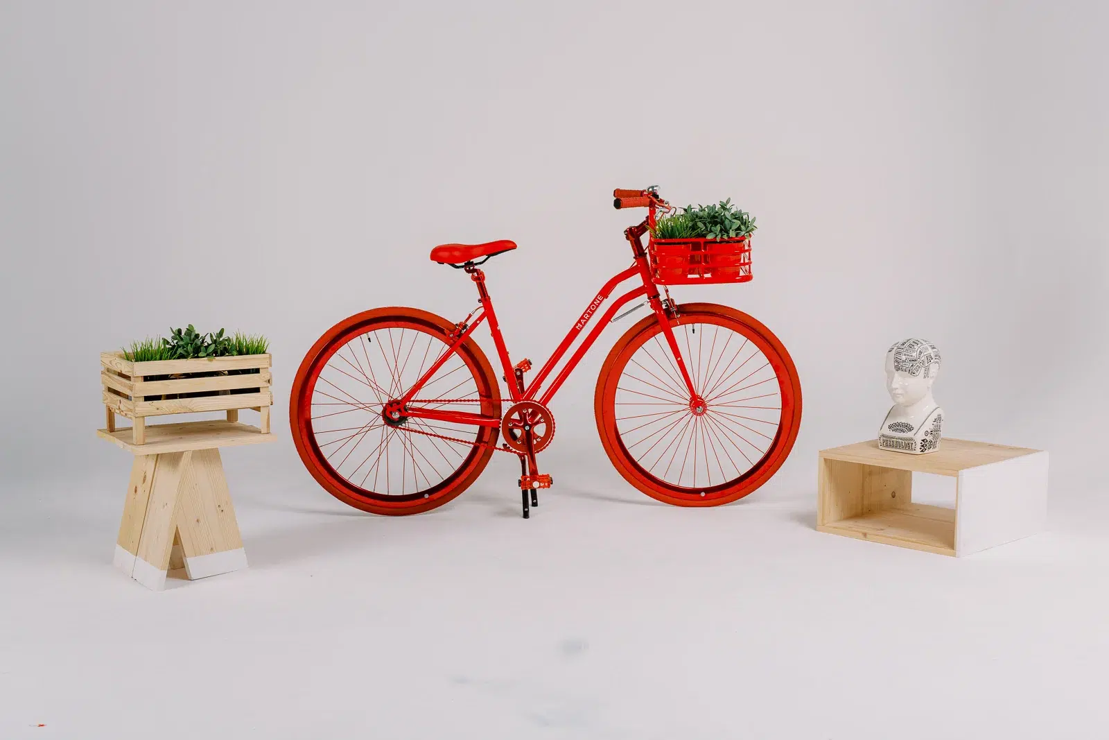 Vélo rouge vif avec panier fleuri, entouré de petits meubles en bois et d'un buste décoratif sur fond blanc.