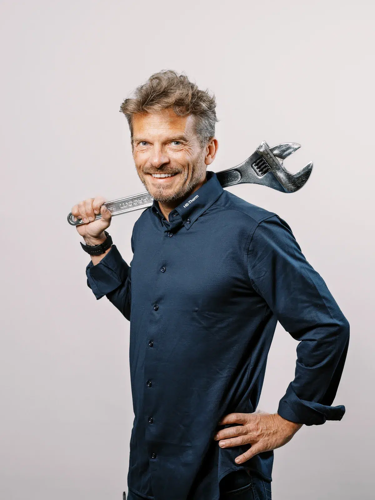 Homme souriant en chemise bleue portant une grande clé à molette sur l'épaule, sur fond clair photographié par un photographe entreprise Lyon.