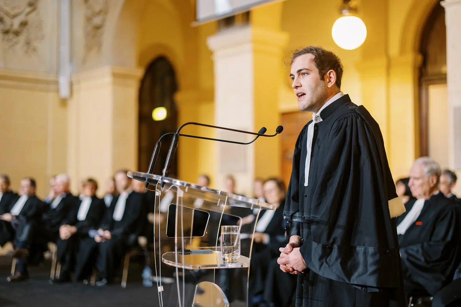 L'intensité d'un concours d'éloquence dans un cadre historique lyonnais. La mise au point précise sur l'orateur au pupitre détache sa silhouette de l'assemblée attentive en arrière-plan.