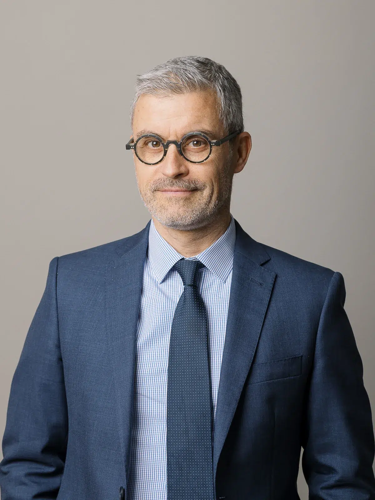 Photographe portrait Lyon : homme en costume bleu et lunettes rondes sur fond gris neutre.