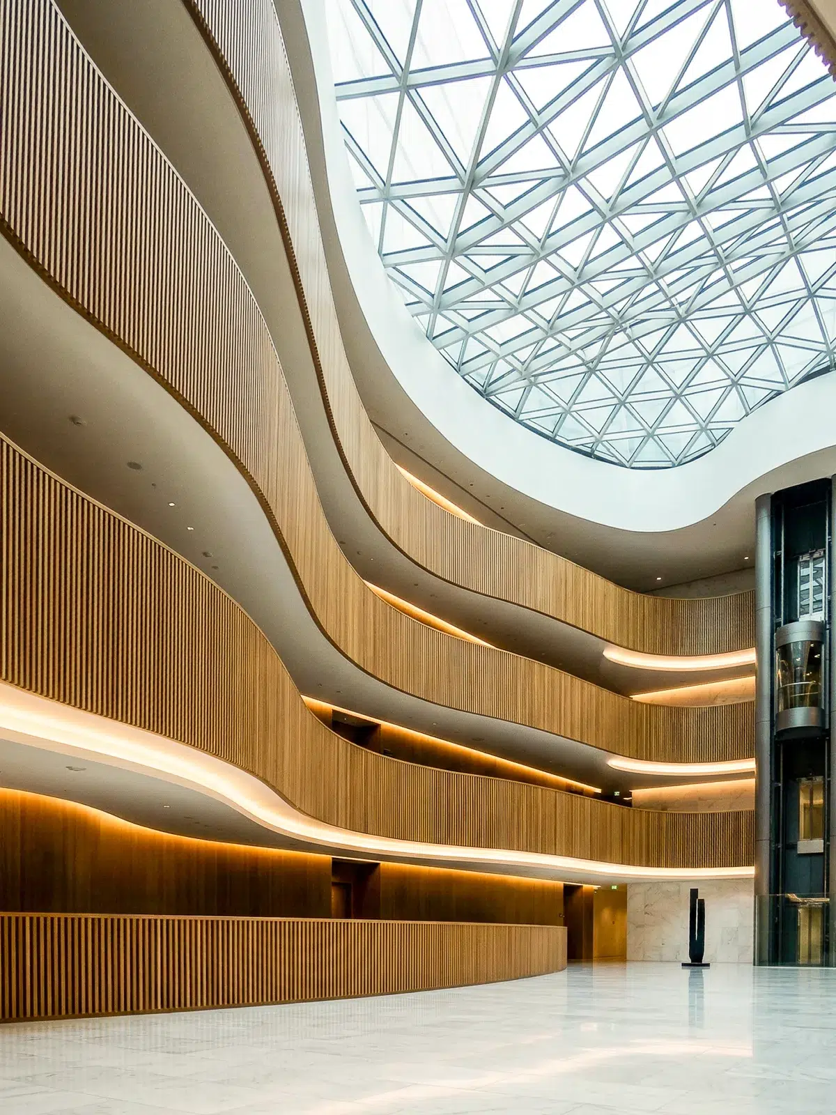 Photographie d'un atrium de grande hauteur. Des balcons aux lignes ondulées, habillés de tasseaux de bois verticaux, créent un rythme visuel dynamique. Une immense coupole géodésique en verre couronne l'espace, inondant le hall de lumière. Un ascenseur panoramique vitré souligne la modernité technique du bâtiment. Le sol en marbre blanc reflète l'ensemble de la structure.