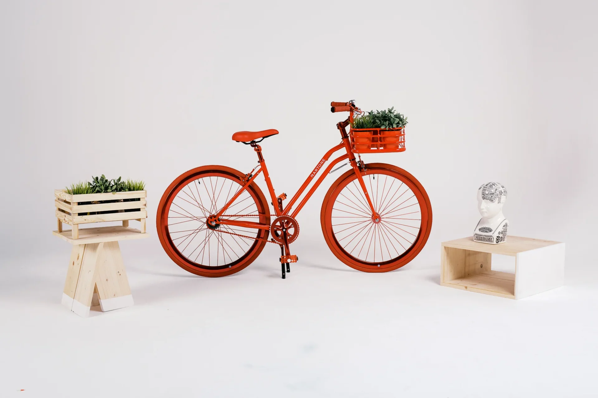 Agence Hula Hoop : vélo rouge Martone, caisses en bois avec plantes et tête de phrénologie sur fond blanc.